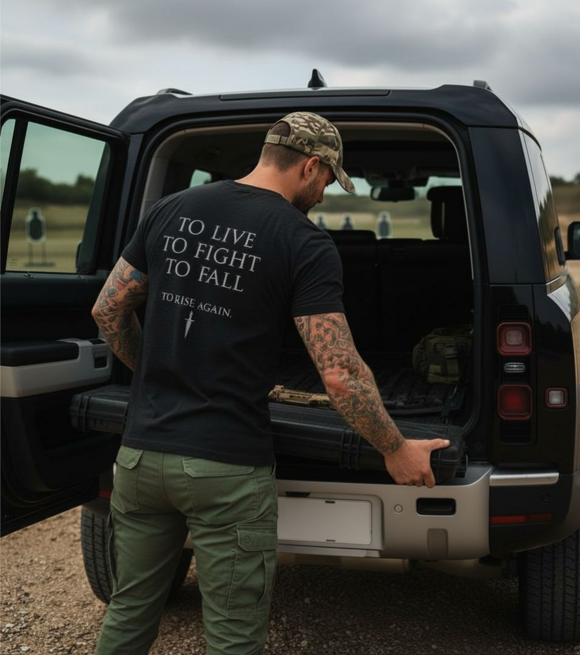 Man with tattoos and a black t-shirt with text standing next to an open SUV.