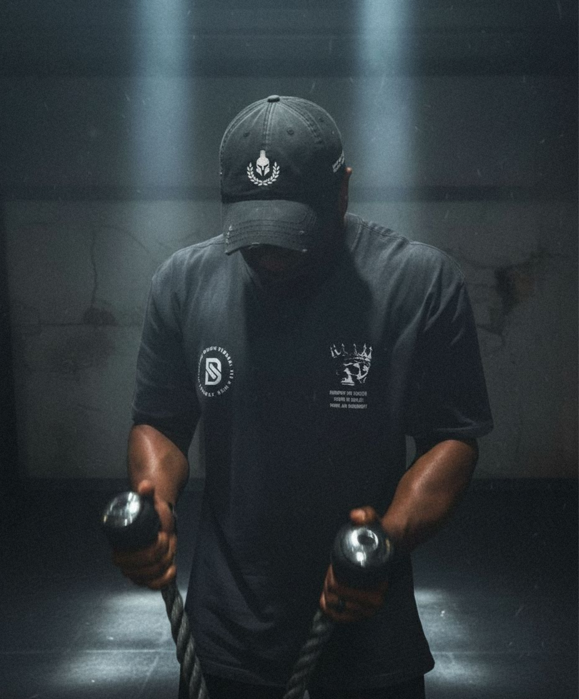 Person wearing a dark cap and t-shirt with logos, holding weights in a dimly lit room.