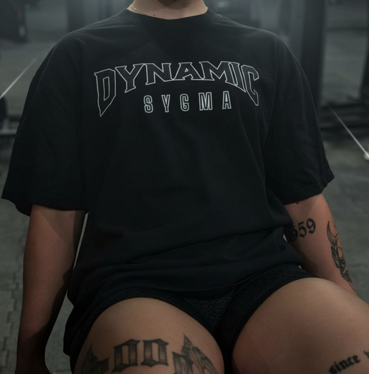 Person wearing a black t-shirt with 'DYNAMIC SYGMA' text, sitting on a bench.