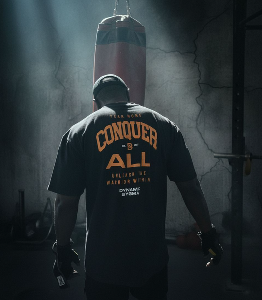 Person wearing a t-shirt with 'Conquer All' text in a dark room with a punching bag.